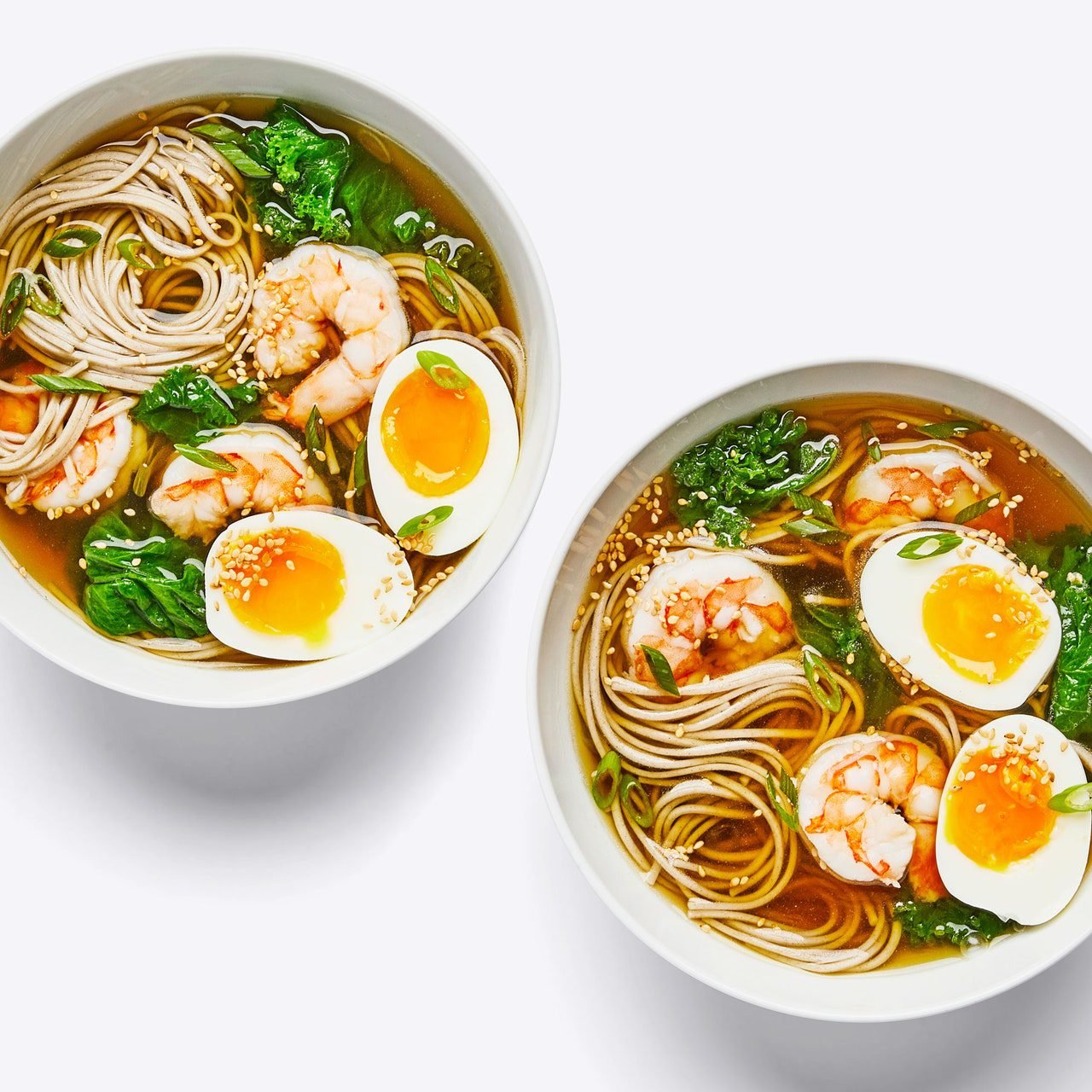 Two bowls of soba soup with shrimp and greens sit on a white tabletop.