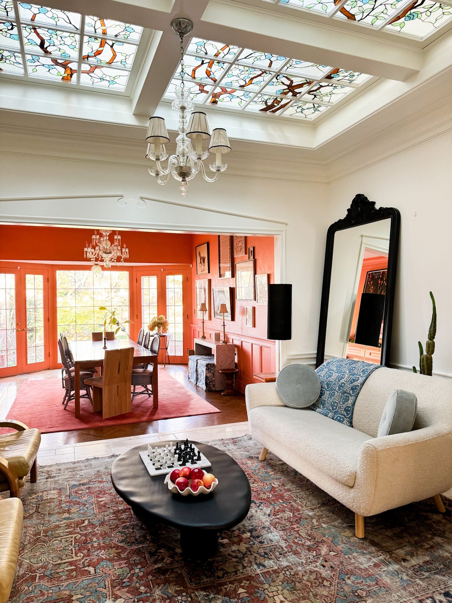 Looking into a sitting room and a dining room beyond it. The sitting room is painted a creamy white color and has a stained-glass ceiling.