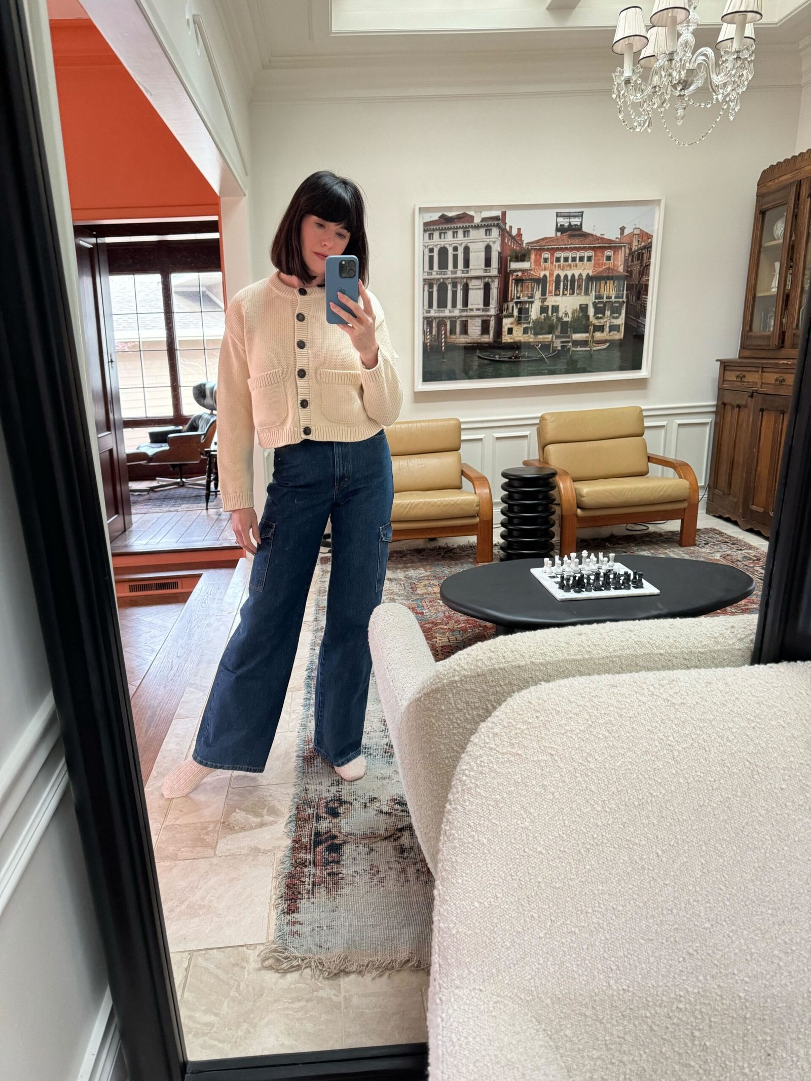 A woman standing in front of a full-length mirror wearing a cream-colored cardigan and dark blue cargo jeans