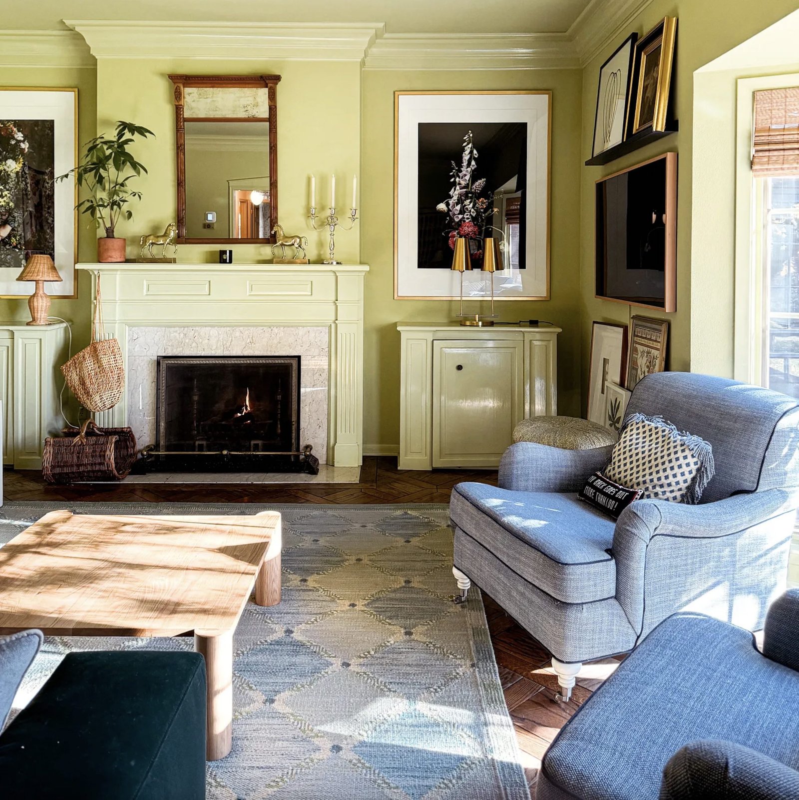 A green living room features a fireplace, two blue chairs, a dark green velvet sofa, an oak square coffee table, and a blue kilim rug.