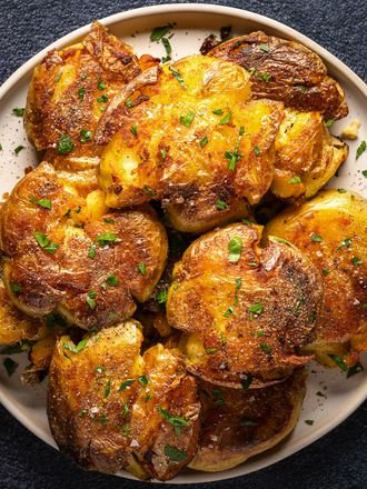 Golden brown crispy smashed potatoes on an oval white plate on a dark textured background.