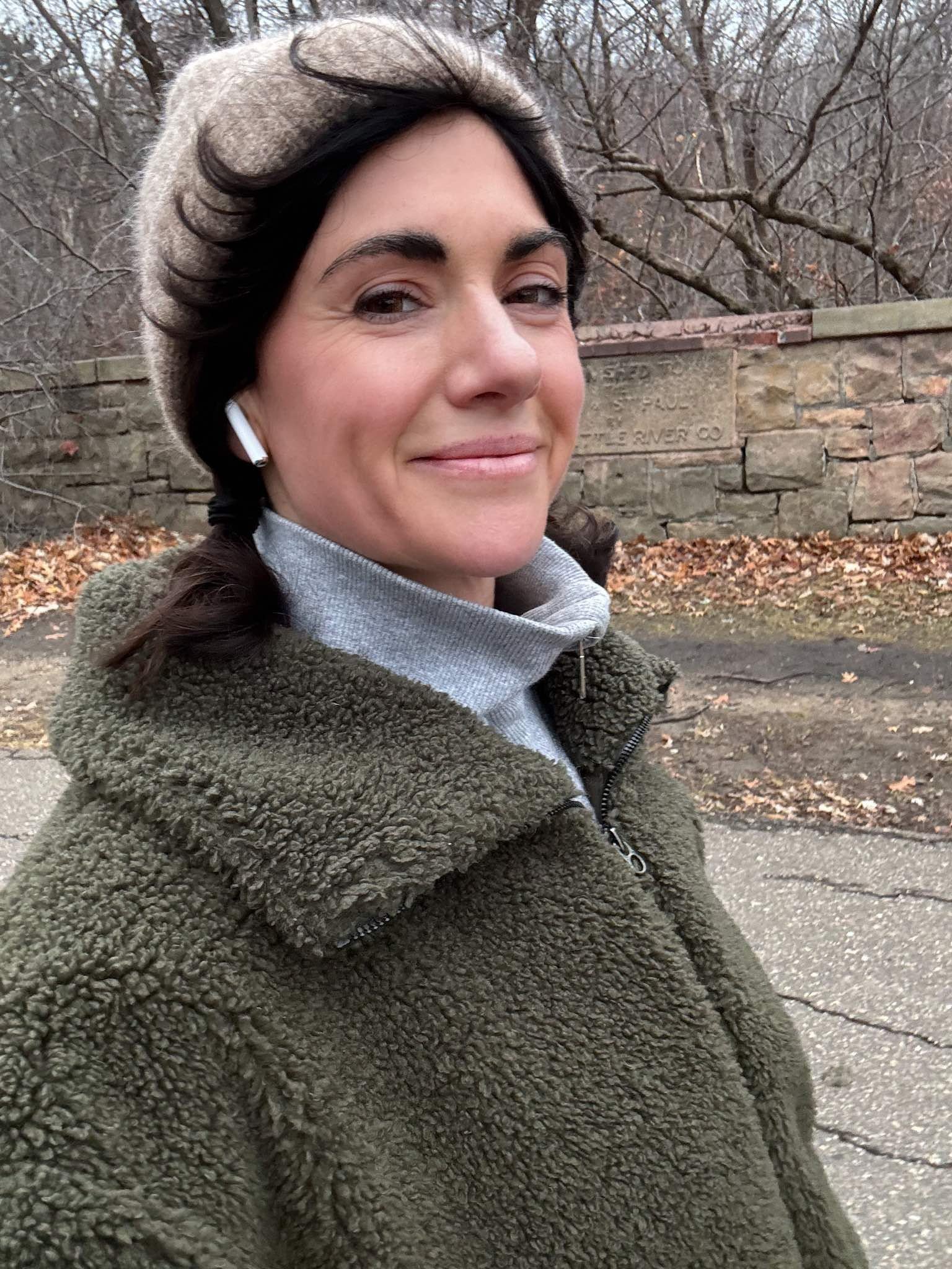 A woman looks into the camera while out for a winter walk, wearing a beige beanie hat, olive green sherpa coat, and gray quarter-zip sweatshirt