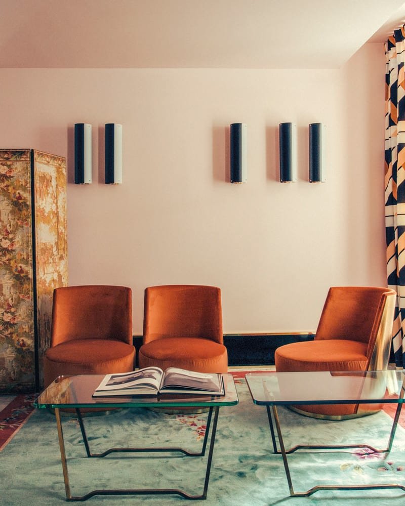 A living space includes a blue and reddish-orange rug, two metal and glass coffee tables, and three orange velvet swivel chairs.